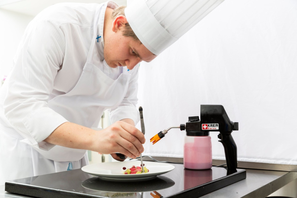Chef plating a gourmet dish with a KREA Swiss electric food sprayer on the commercial kitchen counter